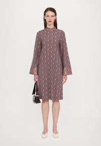 Kurzärmliges, mauvefarbenes Kleid mit geometrischem Muster, das einen hohen Kragen und ausgestellte Ärmel hat. Kombiniert mit einer kleinen schwarzen Handtasche und spitzen Schuhen.