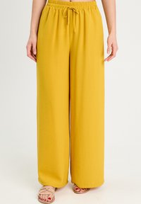 Gelbe weit geschnittene Hosen mit strukturierter Oberfläche, ausgestattet mit einem elastischen Bund und Kordelzug. Kombiniert mit goldenen Sandalen.