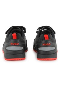 Μαύρα αθλητικά παπούτσια με κόκκινες λεπτομέρειες, που διαθέτουν υφασμένο πάνω μέρος, ανθεκτική σόλα, ανακλαστικές λεπτομέρειες και το όνομα της μάρκας "HUGO" στη φτέρνα.