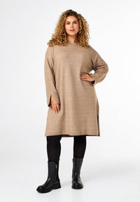 Abito a maglia marrone chiaro con maniche lunghe, fenditure laterali e una vestibilità rilassata, abbinato a stivaletti neri alla caviglia e collant opachi.
