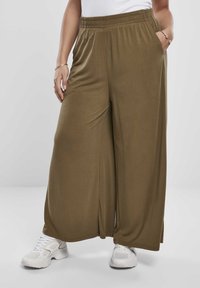 Personne portant un pantalon large vert olive avec taille élastique et baskets blanches, debout les mains dans les poches devant un fond uni.