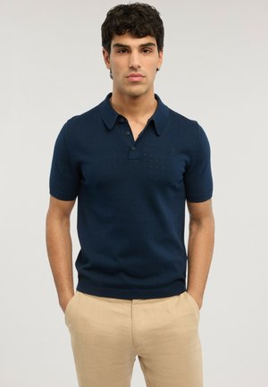 Uomo con capelli scuri e ricci, indossa una polo a maniche corte blu navy e pantaloni beige, in piedi con le mani in tasca su uno sfondo neutro.