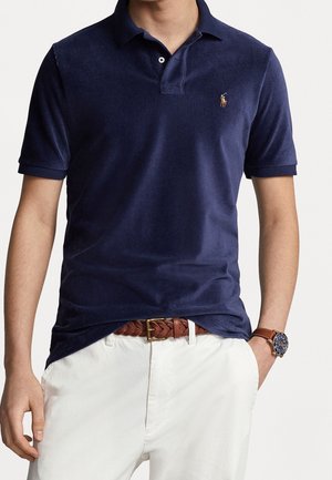 Marineblaues Cord-Polohemd mit klassischem Kragen, kurzen Ärmeln und kleinem gesticktem Logo auf der Brust; kombiniert mit weißen Hosen und einem braunen Gürtel.