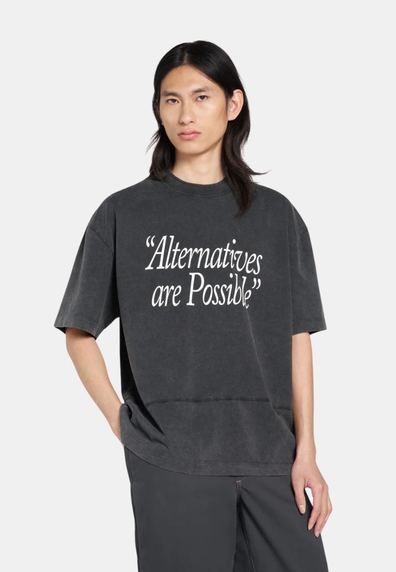 Personne aux longs cheveux noirs portant un t-shirt noir délavé et oversize avec un texte blanc disant "Les alternatives sont possibles" et un pantalon sombre.