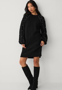 Sort strikkjole med lange, puffede ærmer, der har hvide dekorative perler langs skuldrene. Lavet af strikstof, løst fitting.