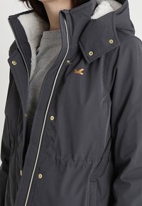 Ciemnoszara kurtka z zamkiem z przodu, podszyta białym polarem, z zatrzaskami na kołnierzu i miedzianym logo na piersi.