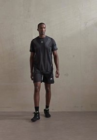 Mann som går innendørs iført svart treningstrøye og shorts med svarte joggesko foran en ensfarget grå vegg.