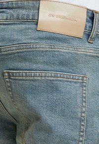 Světle modré džínové kalhoty mají koženou nášivku s nápisem "YZ PREMIUM" na pasu, viditelné švy a jednu zadní kapsu.