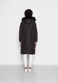 Cappotto puffo nero con cappuccio, dotato di una rifinitura in pelliccia sintetica removibile, texture trapuntata e maniche lunghe, abbinato a leggings bianchi e sneakers.