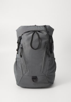 Pelēkā mugursoma, izgatavota no izturīga auduma, ar ruļļu augšdaļu, regulējamiem siksnām un vairākiem kabatām ar rāvējslēdzējiem uzglabāšanai.