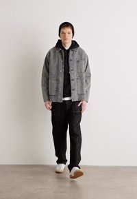 Grå denimjakke med to frontlommer over en svart hettegenser, kombinert med svarte bukser og hvite joggesko. Modellen har på seg en svart lue.