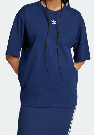 Osoba ubrana w granatowy t-shirt Adidas z białym logo na piersi, dłonie w kieszeniach oraz pasującą granatową spódnicę z białymi paskami.