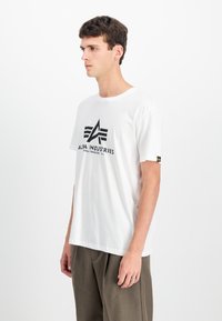 Λευκό βαμβακερό t-shirt με μαύρο γραφικό λογότυπο που περιέχει το κείμενο "ALFA INDUSTRIES", κοντά μανίκια και χαλαρή εφαρμογή.