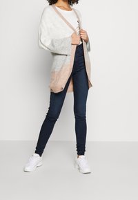 Gilet en tricot blanc, gris et rose pâle avec un design à ouverture sur le devant, des manches larges et une texture douce, associé à un jean skinny noir et des baskets blanches.