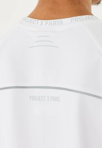 Maglietta sportiva bianca con scollatura tonda, branding "PROJECT X PARIS" in grigio e dettagli a linea curva sul retro.