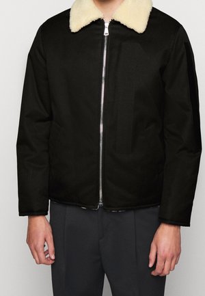 Sort jakke med en blød, cremefarvet shearling-krave, der har en frontlynlås og en glat tekstur. Enkel, struktureret design uden mønstre.