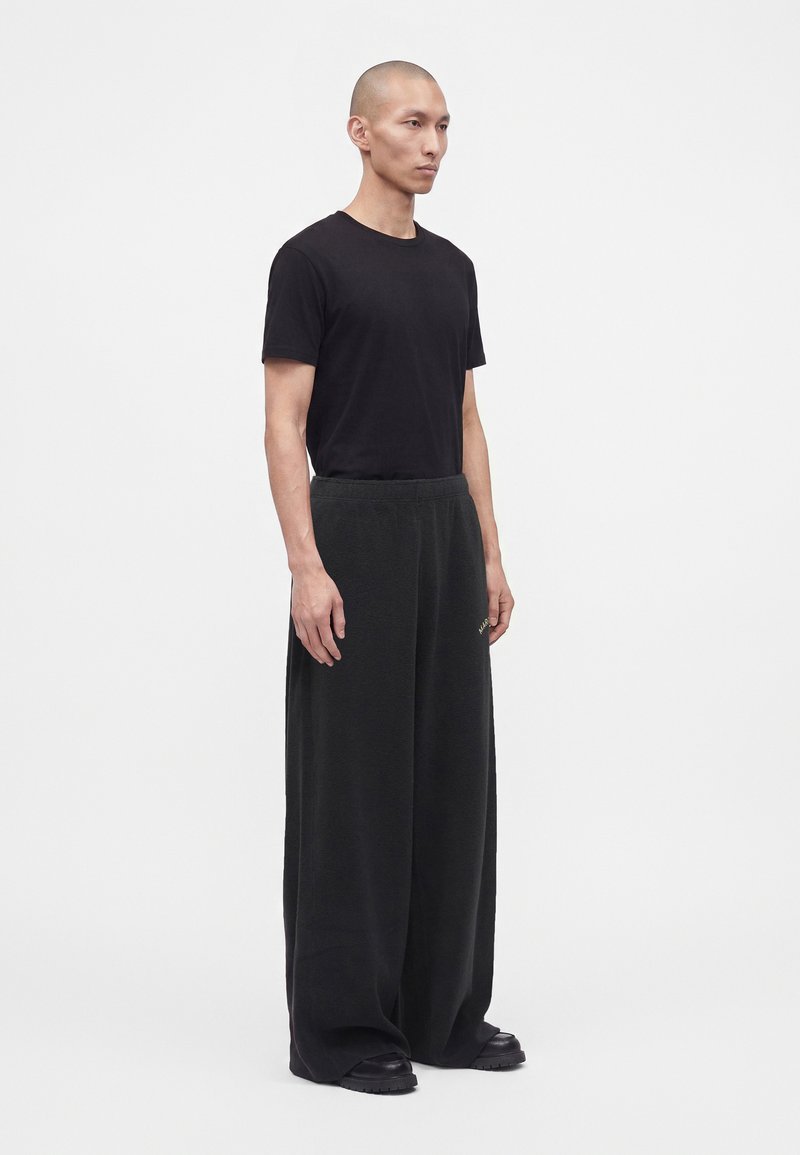 Ein Mann steht mit Blick nach vorne, trägt ein schwarzes T-Shirt und weite schwarze Hosen mit schwarzen Schuhen vor einem einfarbigen weißen Hintergrund.