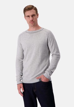 Mann in einem hellgrauen Rundhals-Pullover mit gerippten Bündchen und Saum, kombiniert mit dunkelblauen Jeans, steht mit einer Hand in der Tasche.