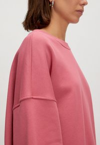 Roze oversized sweatshirt met een ronde halslijn en gedropte mouwen, met een gladde textuur en gestikte accenten op de mouwen.