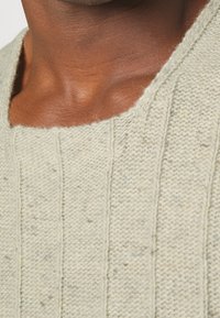 Suéter de punto verde claro con un escote ancho, que presenta una textura de canalé y pequeñas motas de un color más oscuro a lo largo de la tela.