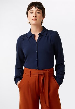 Navy button-up shirt with gathered detailing at shoulders, paired with high-waisted, textured rust-colored trousers featuring a tie belt.