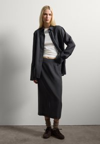 Jonge vrouw staat tegen een witte muur, gekleed in een zwarte lange rok, een zwart open shirt over een witte top, bruin sokken en donkere sneakers.
