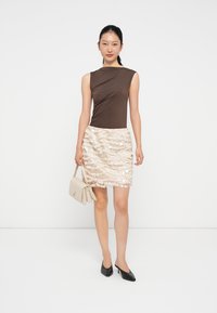 Top sans manches marron avec un design drapé, associé à une jupe beige à sequins avec des motifs ondulés. Chaussures noires à bout pointu et un sac à main blanc.