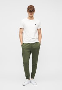 Homme portant un t-shirt blanc, un pantalon de jogging vert olive, des baskets blanches et une casquette marron, debout les mains dans les poches.