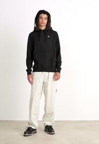 Sudadera negra con capucha, puños elásticos y bolsillo. Pantalones holgados de color claro y zapatos negros con acentos blancos.
