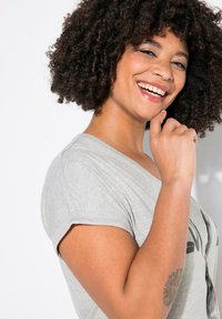 Camiseta de color gris claro con mangas enrolladas, que presenta un diseño gráfico en la parte delantera. Textura suave, corte relajado y escote curvado.