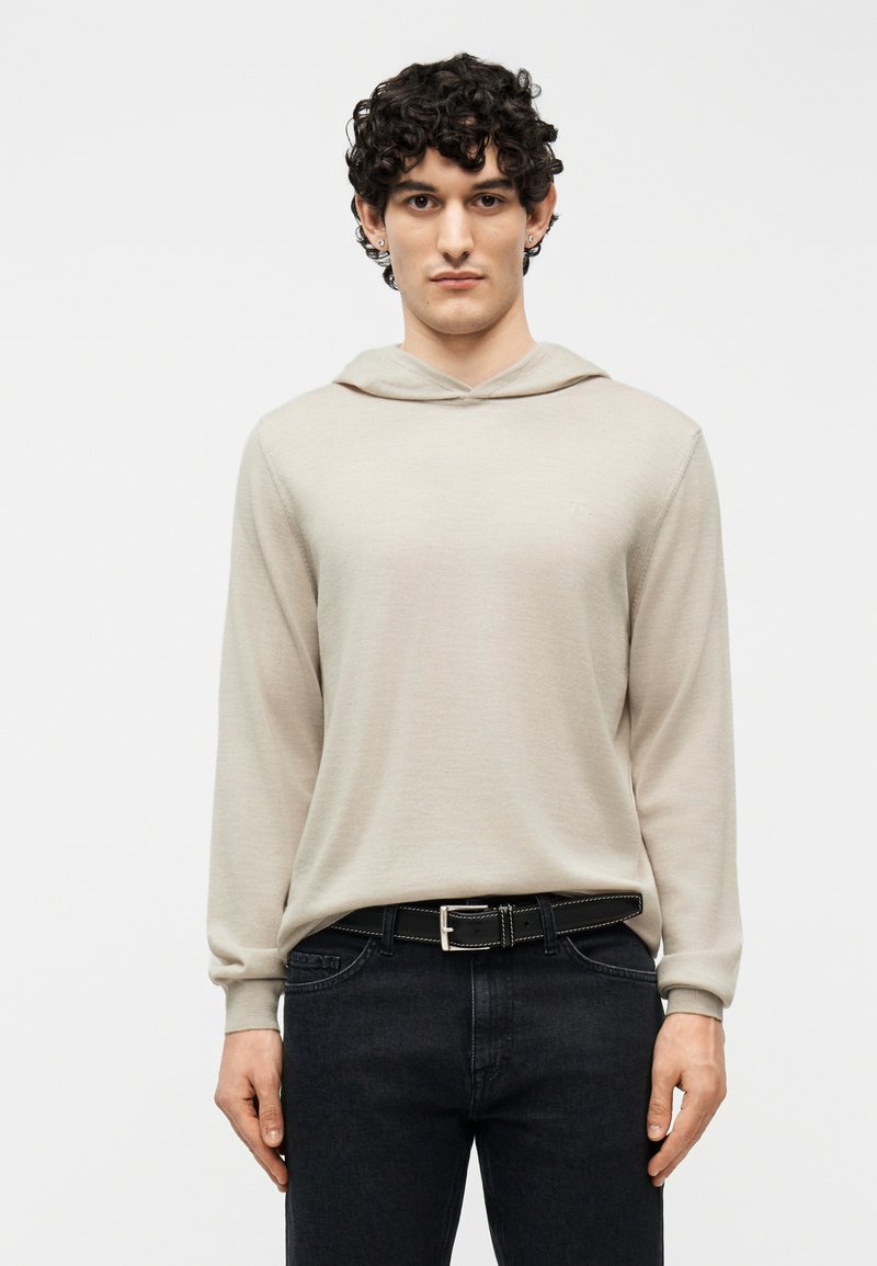 Νεαρός άνδρας με σγουρά μαλλιά, φορώντας μπεζ φούτερ με κουκούλα και μαύρο τζιν με μαύρη ζώνη, στέκεται μπροστά σε απλό λευκό φόντο.