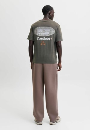 Homme portant un t-shirt graphique vert olive avec le texte "Core Sport" et des étoiles, pantalon marron ample et baskets, debout de dos sur fond blanc.