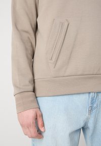 Beige sweatshirt featuring a kangaroo pocket, textured cuffs, and a smooth fabric finish, worn with light blue jeans.