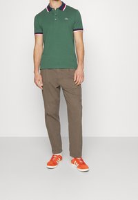 Zelená bavlněná polo košile s bílým a červeným pruhovaným límcem, spárovaná s hnědými kalhotami a oranžovými semišovými teniskami s bílými pruhy.