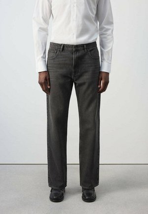 Hombre con vaqueros negros de corte recto, zapatos negros y una camisa blanca de manga larga abotonada, de pie sobre un suelo gris.
