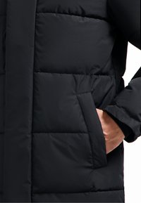 Zwarte pufferjack met een gladde stoftextuur, gestikte details, zijzak en een losse pasvorm. Een deel van de mouw is zichtbaar met een manchet.