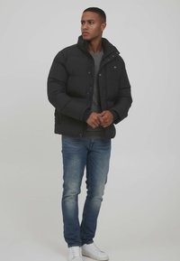 Black puffer jacket with a high collar, button closure, and side pockets. Worn over a gray sweater and blue jeans, with white sneakers.