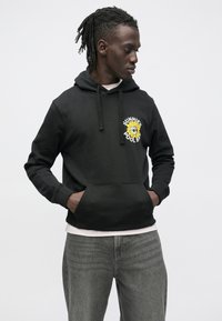 Felpa nera con grafica di un girasole e testo sul petto, con tasca a marsupio e cappuccio con coulisse regolabile, abbinata a jeans grigi.