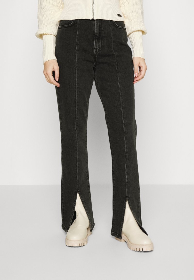 Black denim pants with front slits, high waist, and straight leg cut. Features contrast stitching and side pockets. Paired with beige ankle boots.
