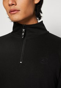 Sort lynlås fleece med høj krave. Har en halv lynlås, broderet logo og glat tekstur. Enkel, minimalistisk design.
