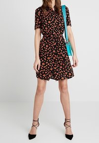 Svart kortärmad klänning med röd blommönster, bälte i midjan, tillsammans med en turkos handväska och svarta remsandaler.