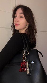 Sort tekstureret håndtaske med to håndtag, med en guldfarvet nøglekæde med en rød bærsmykke og metalmærke. Enkel, struktureret design.