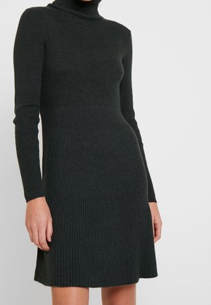 Femme portant une robe noire moulante en tricot côtelé à manches longues avec un col haut, sur un fond clair uni.