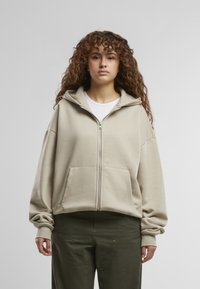 Beige zip-up hoodie met een capuchon, in een relaxed fit, met voorzakken en geribbelde manchetten. Gedragen over een wit T-shirt en gecombineerd met groene broek.