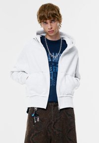 Szürke cipzáros kapucnis pulóver, bélelt kapucnival, sötét kék grafikás pólóra húzva, barna, paisley mintás nadrággal párosítva.