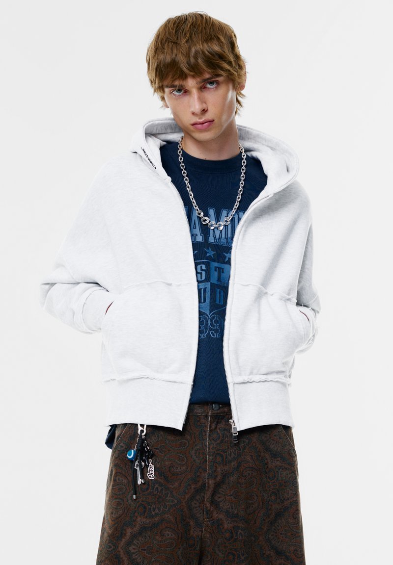 Szürke cipzáros kapucnis pulóver, bélelt kapucnival, sötét kék grafikás pólóra húzva, barna, paisley mintás nadrággal párosítva.