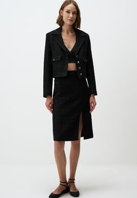 Veste noire texturée au design court, dotée de deux poches avant, associée à une jupe noire à fente haute, offrant une finition lisse et une silhouette près du corps.