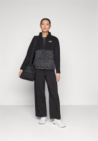 Black fleece pullover with a half-zip and a textured, patterned lower section, paired with black wide-leg trousers and white sneakers.