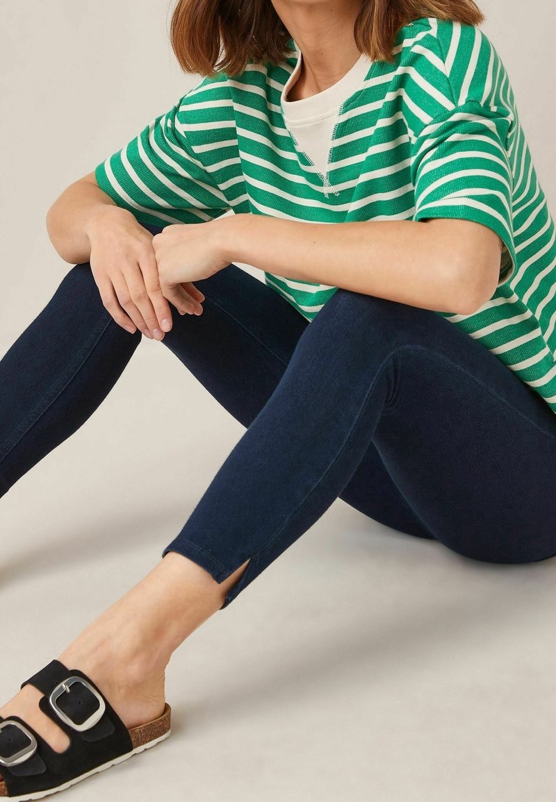 Femme assise en tailleur portant une chemise rayée verte et blanche, un pantalon skinny bleu foncé avec des fentes aux chevilles, et des sandales noires à boucle.