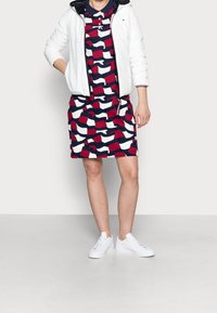 Weiße Daunenjacke über einem gemusterten Kleid in Rot, Weiß und Marineblau. Das Modell trägt weiße Sneakers. Glattes Material, lässiges Design und kurze Ärmel.
